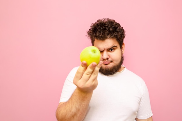Gracioso joven con sobrepeso parado sobre un fondo rosa se ve sorprendido por la manzana en su mano