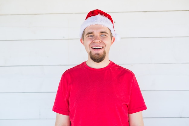 Gracioso joven con sombrero de santa y camisa sonriendo sobre la pared blanca