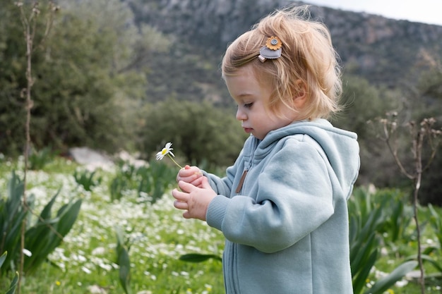 Gracioso lindo adorable caucásica rubia bebé niña pequeña en campo verde de manzanilla margarita oliendo floresNiño curiosoniño infantilambiente de aprendizajenaturalezamontañas en primavera o verano