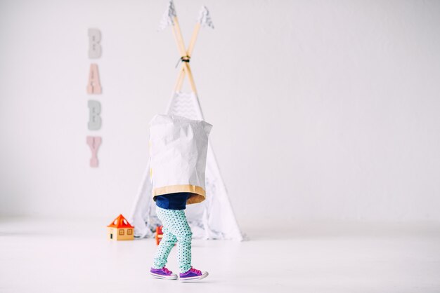 Gracioso niño corriendo por la habitación luminosa con una bolsa de papel en la cabeza en wigwam para niños