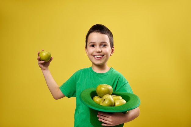 Foto gracioso niño se encuentra aislado en la superficie amarilla y sostiene un sombrero de duende irlandés lleno de manzanas en una mano y una manzana verde en la otra