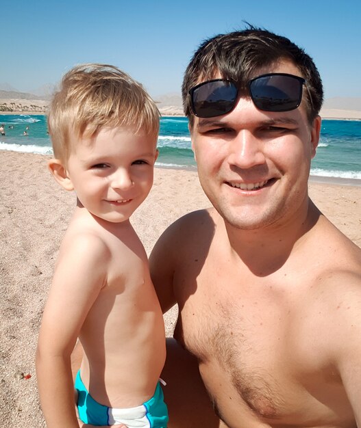 Gracioso retrato de joven padre haciendo imagen selfie con su hijo pequeño en la playa del mar en el sol brillante. Familia relajándose y pasando un buen rato durante las vacaciones de verano.