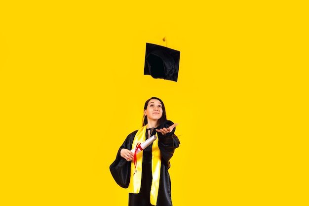 Graduada con un diploma, levantando su gorro de graduación en el aire y celebrando sobre una pared amarilla