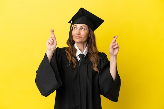 Graduado de meia-idade isolado em um fundo amarelo com os dedos se cruzando