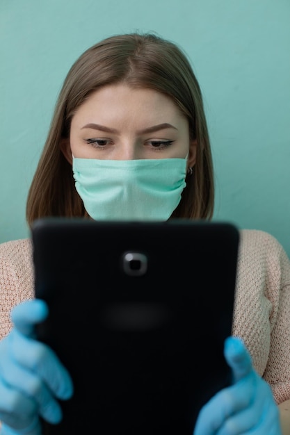 Un graduado de la escuela secundaria sentado en una cama con una tableta en la mano en guantes de goma y una máscara protectora