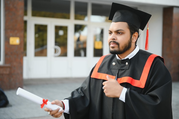 Graduado indio en bata de graduación con diploma en espacio de copia del campus universitario