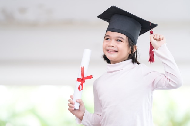 Graduado de niño de escuela asiática femenina con un gorro de graduación tiene un papel de certificado enrollado