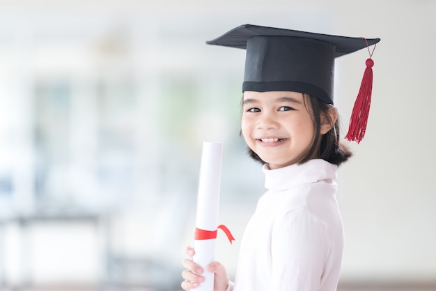 Graduado de niño de escuela asiática femenina con un gorro de graduación tiene un papel de certificado enrollado