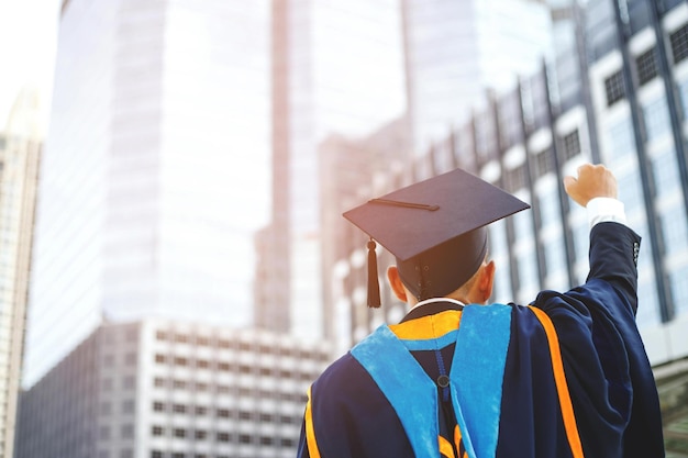 Un graduado recibió un disparo en la parte posterior del joven estudiante en la mano levantando los brazos sobre el puño y el pulgar hacia arriba durante el éxito de la graduación de la universidad Concepto educación felicitación