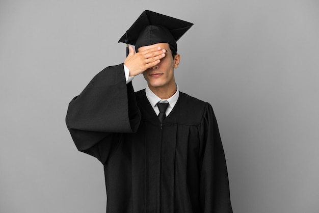 Graduado universitario argentino joven aislado sobre fondo gris cubriendo los ojos con las manos. No quiero ver algo