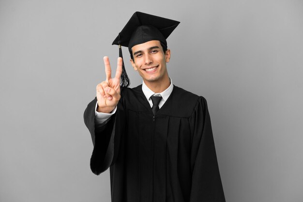 Graduado universitario argentino joven aislado sobre fondo gris sonriendo y mostrando el signo de la victoria