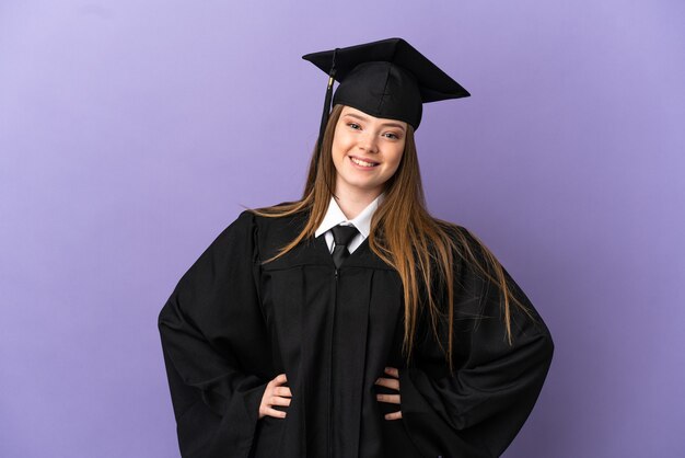 Graduado universitario joven sobre fondo púrpura aislado posando con los brazos en la cadera y sonriendo