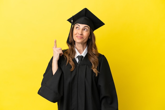 Graduado universitario de mediana edad aislado sobre fondo amarillo apuntando hacia arriba y sorprendido