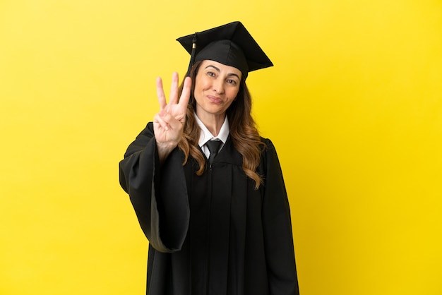 Graduado universitario de mediana edad aislado sobre fondo amarillo feliz y contando tres con los dedos