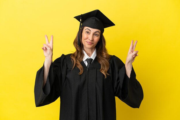 Graduado universitario de mediana edad aislado sobre fondo amarillo que muestra el signo de la victoria con ambas manos