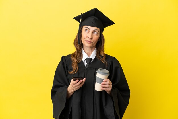 Graduado universitario de mediana edad aislado sobre fondo amarillo sosteniendo café para llevar y un móvil mientras piensa en algo