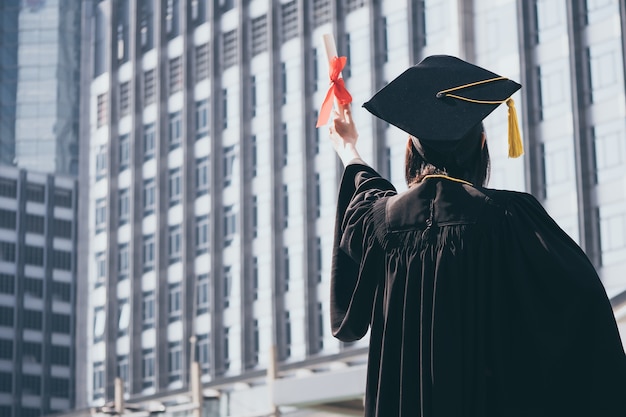 Graduierungstag, hintere Ansicht der Asiatin mit der Staffelungskappe und -kleid, die Diplom halten