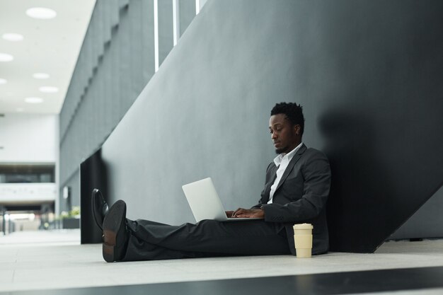 Gráfico retrato de cuerpo entero del empresario afroamericano con laptop mientras está sentado en el piso en el pasillo de la oficina, espacio de copia