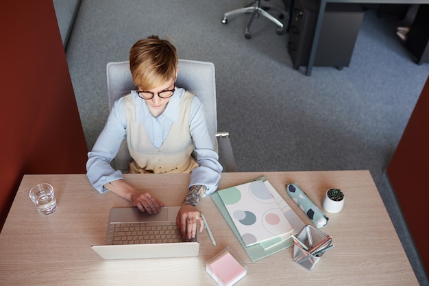 Grafische Ansicht von oben nach unten an der modernen tätowierten Geschäftsfrau unter Verwendung des Laptops am Schreibtisch während der Arbeit im Büro, Kopierraum