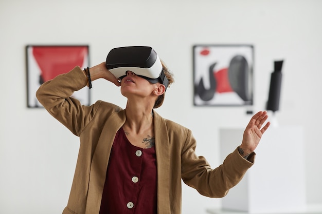 Grafisches Taillen-Hochporträt der modernen jungen Frau, die VR-Ausrüstung trägt, während immersive Erfahrung an Kunstausstellungsausstellung genießt