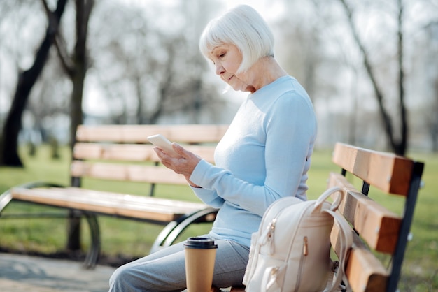 Gran artilugio. Mujer de edad concentrada escribiendo en su teléfono mientras está sentado en el banco