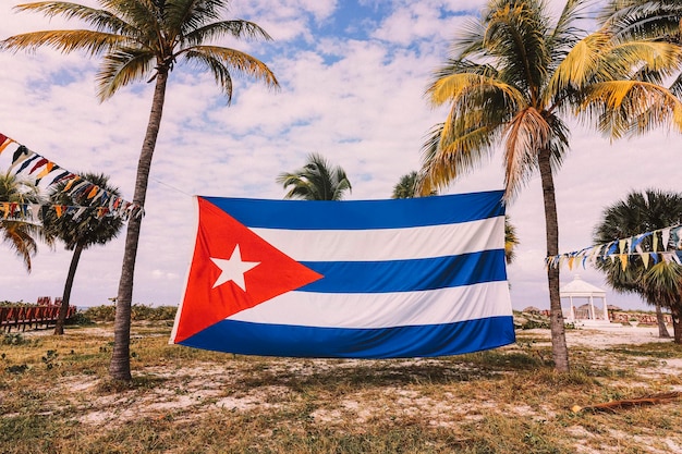 Gran bandera de Cuba está colgada entre palmeras en la playa del mar Caribe contra un cielo nublado Hermoso paisaje tropical en el fondo