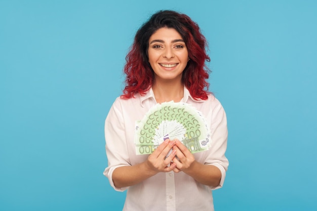 Gran beneficio, puesta en marcha exitosa. Retrato de una mujer feliz y sonriente con el pelo rojo elegante que sostiene billetes en euros, muestra dinero y alardea de altos ingresos. Foto de estudio interior aislado sobre fondo azul.