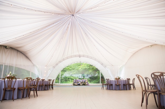 Gran carpa de boda blanca con hermosas decoraciones. Mesas con adornos florales y sillas de madera.