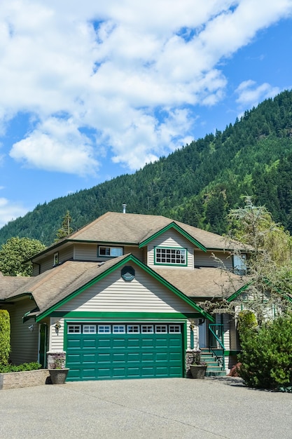 Gran casa familiar con amplio camino de hormigón, amplia puerta de garaje verde decorada en el patio delantero Casa residencial con fondo de cielo azul y montaña