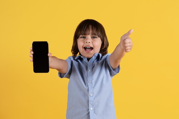 Gran contenido para niños Un niño pequeño emocionado que muestra un teléfono inteligente con pantalla en blanco a la cámara y gesticula con el pulgar hacia arriba
