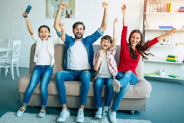 Un gran equipo familiar emocionado viendo un partido de fútbol en la televisión mientras se sientan juntos en el sofá de casa