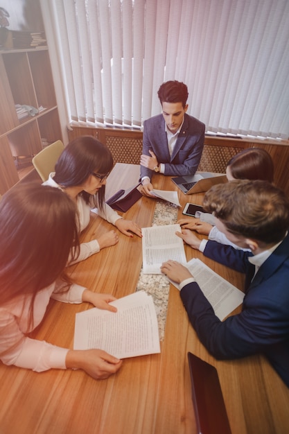 Un gran equipo de personas está trabajando en una mesa para computadoras portátiles, tabletas y papeles.