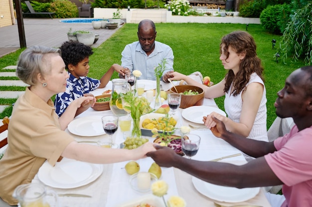 Gran familia intercultural de tres generaciones tomadas de la mano durante la oración mientras están sentadas junto a una mesa servida con comida y bebidas caseras