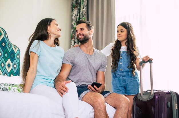 Foto gran familia de turistas felices y emocionados están usando un teléfono inteligente mientras están sentados en la cama en la habitación del hotel