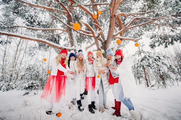 Un gran grupo de niñas con mandarinas están de pie en el bosque de invierno.Niñas en ropa roja y blanca con frutas en un bosque cubierto de nieve