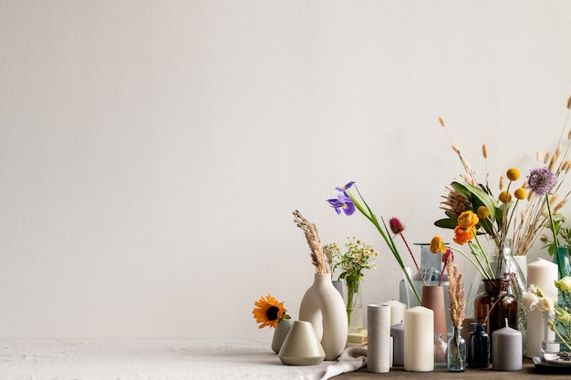 Gran grupo de varios jarrones de cerámica, arcilla y vidrio hechos a mano creativos con flores frescas y secas y velas aromáticas en la mesa junto a la pared