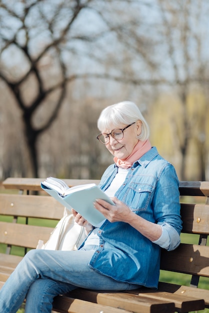 Gran libro. Mujer rubia alegre leyendo un libro mientras está sentado en el banco