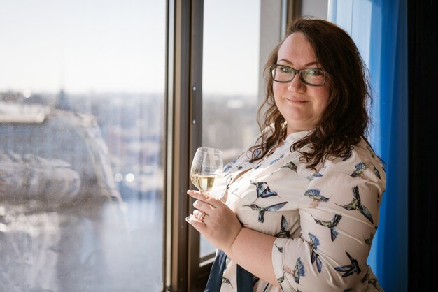 Gran mujer de pie en la ventana con una copa de vino, el concepto de soledad