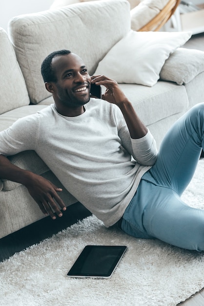 ¡Una gran noticia! Apuesto joven africano hablando por teléfono y sonriendo mientras está sentado en la alfombra en casa