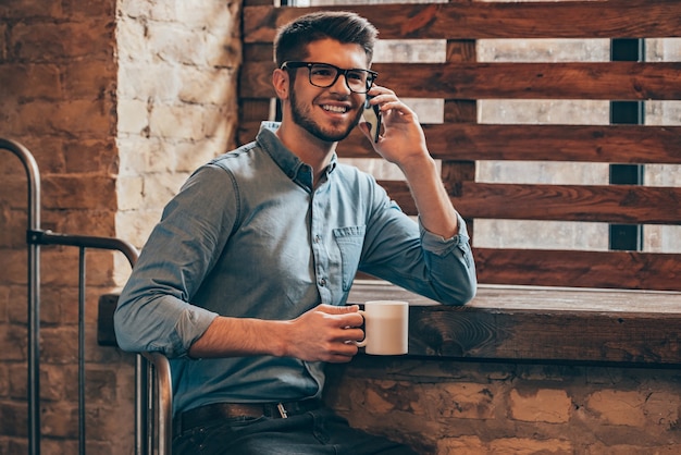 ¡Una gran noticia! Apuesto joven hablando por teléfono móvil y sonriendo mientras está sentado junto a la ventana en el interior del desván con una taza de café en la mano