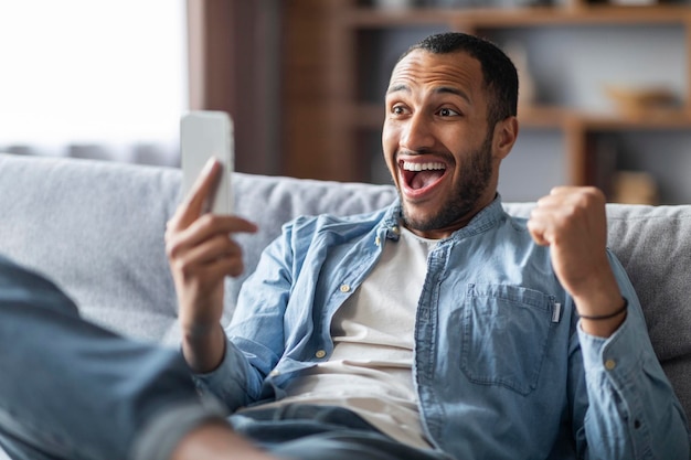Gran noticia Hombre negro encantado mirando la pantalla del teléfono inteligente y celebrando el éxito