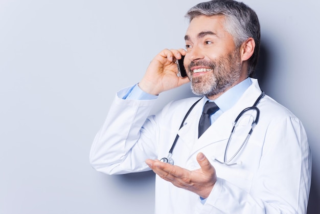 Foto ¡una gran noticia! médico de cabello gris maduro feliz hablando por teléfono móvil y gesticulando mientras está de pie contra el fondo gris