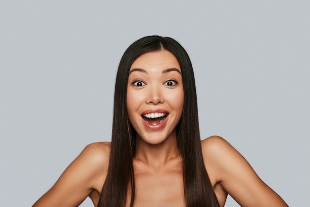 ¡Una gran noticia! Mujer asiática joven sorprendida mirando a la cámara y sonriendo mientras está de pie contra el fondo gris