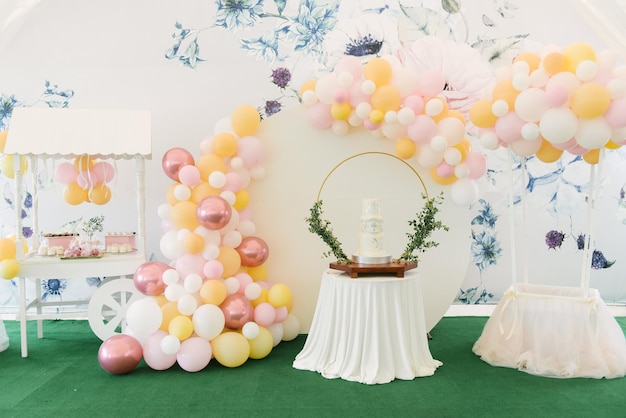 Un gran pastel blanco de tres niveles para el bautismo de un niño con ramitas verdes en el fondo de una hermosa zona fotográfica con globos y una barra de dulces