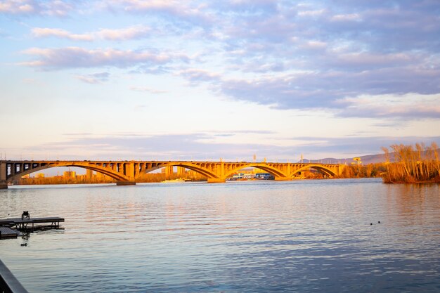 Gran puente en los rayos del sol poniente sobre el río. Cielo nublado. Puente comunal, río Yenisei, Krasnoyarsk, Siberia, Rusia.