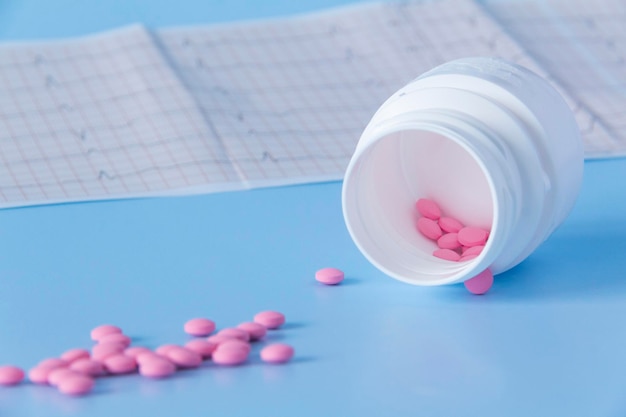 Un gran puñado de pastillas rosadas se derramó de un frasco blanco sobre un electrocardiograma del corazón sobre un fondo azul El concepto de un estilo de vida saludable y un examen médico oportuno