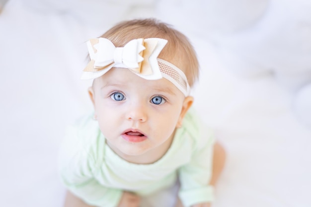 Un gran retrato de una linda niñita con grandes ojos azules mirando a la cámara con un lazo en la cabeza en una cuna con un traje de algodón verde