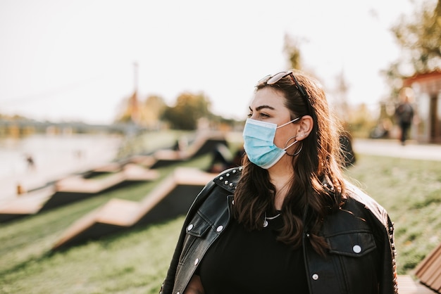 Gran retrato de una mujer hermosa en una máscara médica y chaqueta de cuero en un parque en la naturaleza 1