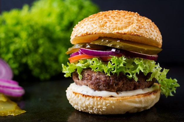 Foto gran sándwich - hamburguesa con carne de vacuno, encurtidos, tomate y salsa tártara sobre fondo negro.