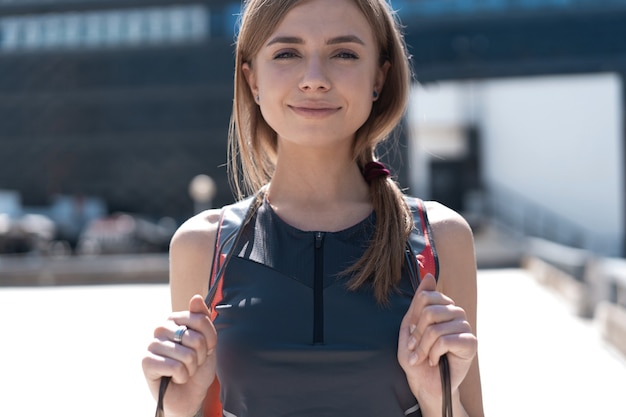 ¡Gran trabajo! Feliz joven deporte en elegante traje de entrenamiento con saltar la cuerda sobre sus hombros está sonriendo, de pie afuera